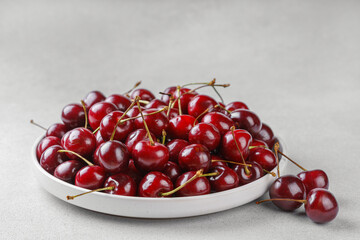 Heap of fresh ripe red sweet cherry with stems in white plate on light textured background. Natural summer fruit, healthy organic food, harvest and seasonal concepts.