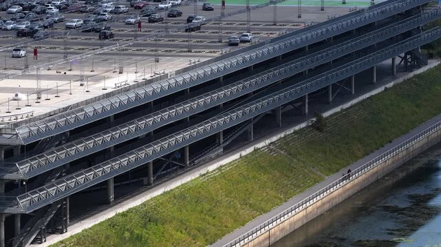 Elevated view of a modern multi-tier parking facility with metal ramps and a riverside promenade featuring cyclists and urban greenery.