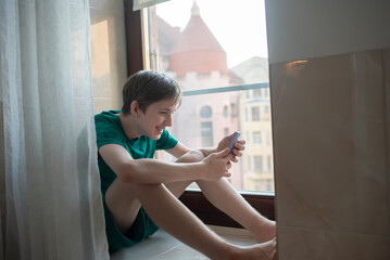 Happy 12 year old boy in pajamas sitting on windowsill at home, looking at smartphone.