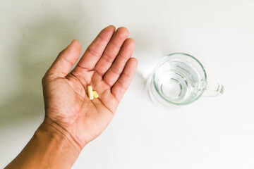 A hand holding yellow tablets with a glass of water on a white background.