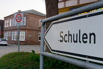 Obraz premium A white arrow on metal poles points left with German school signage. In the background, a traffic sign displays Zone 30 indicating a 30 km h speed limit area. The setup marks a school zone.