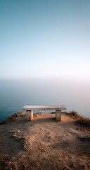 A solitary weathered stone bench sits atop a cliff overlooking a calm, misty ocean under a pale sky