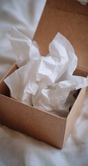 An open, square, kraft paper gift box sits on a white fabric background; crumpled white tissue paper fills its interior