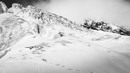 A high-contrast black and white image of a glacier’s steep snowy surface and jagged ice formations, with a mist-covered mountain range in the background creating a sense of scale and mystery.