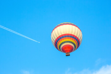 Colorful hot air balloon soaring high above clear blue sky with jet plane trail