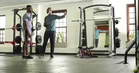 African American men entering gym discussing workout plan on tablet by squat rack before exiting