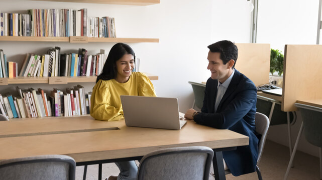 Indian and Hispanic teammates sitting at table in modern coworking office with laptop, discussing joint task or project details. Positive sales manager consulting company client, showing presentation