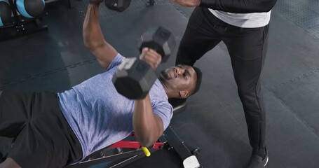 African American personal trainer guiding client bench pressing with dumbbells at gym for safety - Powered by Adobe