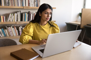 Focused Indian businesswoman working on laptop in workplace, editing document, assessing detailed proposals, contracts, or partnership opportunity, respond on business e-mails, brainstorming solutions