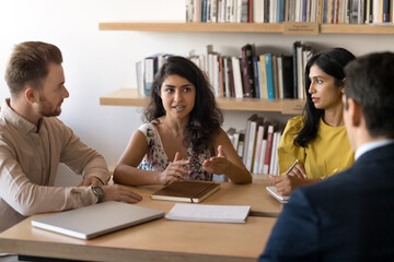 Fototapeta premium Indian woman, team leader make speech during briefing with multiethnic colleagues, listening her attentively, sit together at desk in boardroom, solve work-related matters, share thoughts and opinions