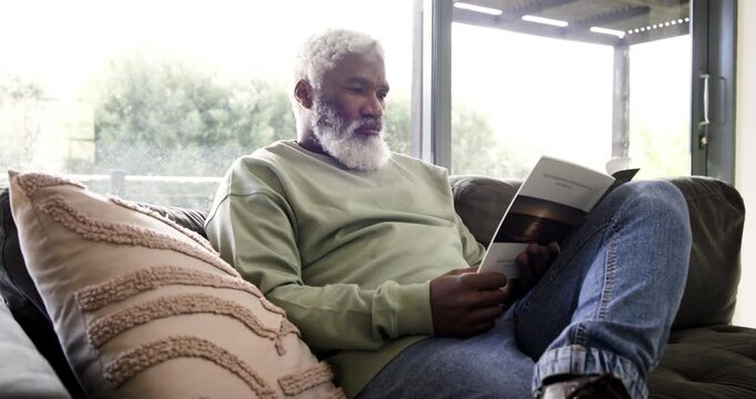 Senior African American man settling on sofa opening brochure and reading intently by window