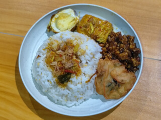 A dish of mixed rice with side dishes of orek tempeh, salted egg, perkedel, and yellow spiced egg, served on a white melamine plate on a wooden table. Traditional Indonesian food mixed rice.