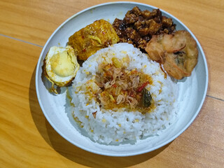 A dish of mixed rice with side dishes of orek tempeh, salted egg, perkedel, and yellow spiced egg, served on a white melamine plate on a wooden table. Traditional Indonesian food mixed rice.