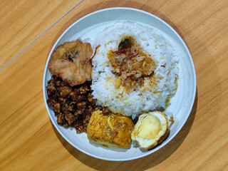 A dish of mixed rice with side dishes of orek tempeh, salted egg, perkedel, and yellow spiced egg, served on a white melamine plate on a wooden table. Traditional Indonesian food mixed rice.