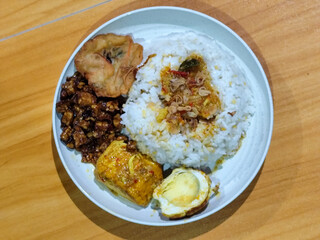 A dish of mixed rice with side dishes of orek tempeh, salted egg, perkedel, and yellow spiced egg, served on a white melamine plate on a wooden table. Traditional Indonesian food mixed rice.