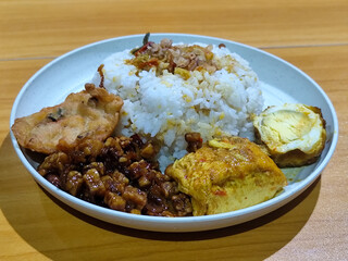 A dish of mixed rice with side dishes of orek tempeh, salted egg, perkedel, and yellow spiced egg, served on a white melamine plate on a wooden table. Traditional Indonesian food mixed rice.