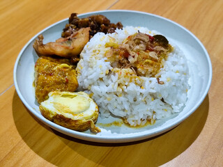 A dish of mixed rice with side dishes of orek tempeh, salted egg, perkedel, and yellow spiced egg, served on a white melamine plate on a wooden table. Traditional Indonesian food mixed rice.