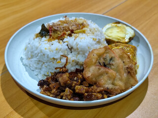 A dish of mixed rice with side dishes of orek tempeh, salted egg, perkedel, and yellow spiced egg, served on a white melamine plate on a wooden table. Traditional Indonesian food mixed rice.