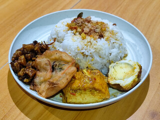 A dish of mixed rice with side dishes of orek tempeh, salted egg, perkedel, and yellow spiced egg, served on a white melamine plate on a wooden table. Traditional Indonesian food mixed rice.