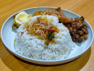 A dish of mixed rice with side dishes of orek tempeh, salted egg, perkedel, and yellow spiced egg, served on a white melamine plate on a wooden table. Traditional Indonesian food mixed rice.