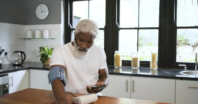 Senior African American man tapping smartphone, starting BP monitor on countertop for health check