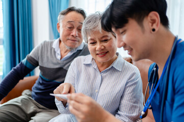 asian senior woman using digital thermometer measure temperature instructed by physiotherapist. home caregiver nurse visit retirement patient for health check up recovery process medical service