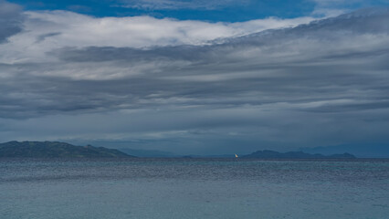 A serene seascape. Turquoise ocean. A lonely white sail on the horizon. Silhouettes of mountains against the blue sky and clouds. Madagascar.