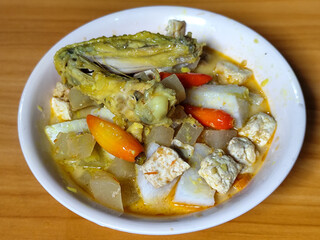 Traditional Indonesian vegetable lontong dish. Lontong opor ayam with sliced chayote, carrots, tofu and sliced red chilies, served in a white ceramic bowl on a wooden table.