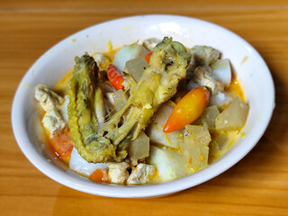 Traditional Indonesian vegetable lontong dish. Lontong opor ayam with sliced chayote, carrots, tofu and sliced red chilies, served in a white ceramic bowl on a wooden table.