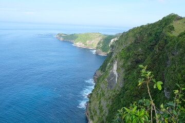 Fototapeta premium Lush green cliffs drop steeply into the blue ocean along the rugged coastline of Kelingking Beach, Nusa Penida, Bali.