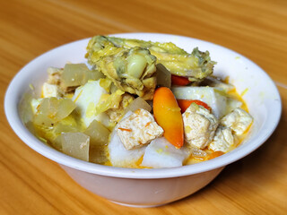 Traditional Indonesian vegetable lontong dish. Lontong opor ayam with sliced chayote, carrots, tofu and sliced red chilies, served in a white ceramic bowl on a wooden table.
