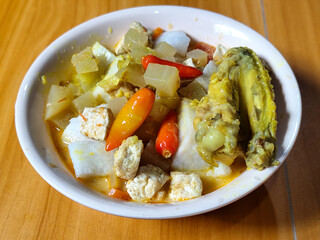 Traditional Indonesian vegetable lontong dish. Lontong opor ayam with sliced chayote, carrots, tofu and sliced red chilies, served in a white ceramic bowl on a wooden table.