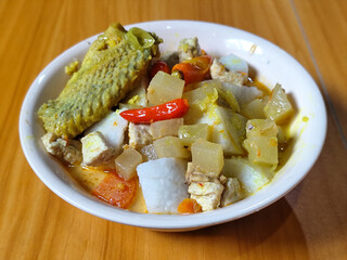 Traditional Indonesian vegetable lontong dish. Lontong opor ayam with sliced chayote, carrots, tofu and sliced red chilies, served in a white ceramic bowl on a wooden table.
