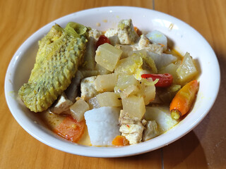 Traditional Indonesian vegetable lontong dish. Lontong opor ayam with sliced chayote, carrots, tofu and sliced red chilies, served in a white ceramic bowl on a wooden table.