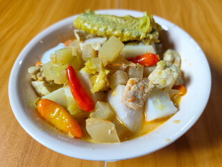 Traditional Indonesian vegetable lontong dish. Lontong opor ayam with sliced chayote, carrots, tofu and sliced red chilies, served in a white ceramic bowl on a wooden table.