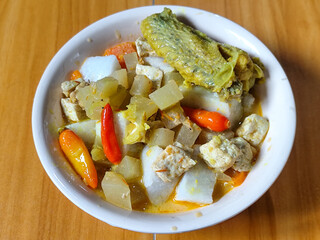 Traditional Indonesian vegetable lontong dish. Lontong opor ayam with sliced chayote, carrots, tofu and sliced red chilies, served in a white ceramic bowl on a wooden table.