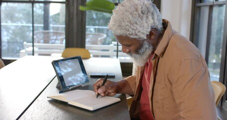After notification senior African American man writing in notebook at home and viewing tablet - Powered by Adobe