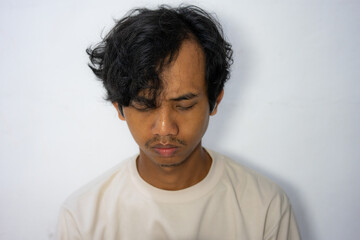 A young Indonesian-asian man with a serious expression and messy hair, standing against a white wall. Perfect for portraying emotions, portraits, or lifestyle projects.