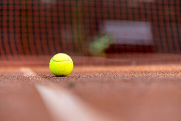 Tennis Ball on Clay Court with Net in Background Close-up of a bright yellow tennis ball resting on the baseline of a clay tennis court, with the net softly blurred in the background. 