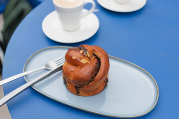 A poppy-flavored brioche decorated with almond petals on a beautiful oval ceramic plate with cappuccino coffee in elegant white cups on a blue table.