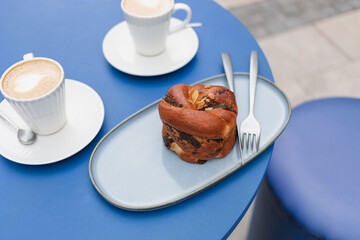 Poppy-flavored brioche with cappuccino coffee in an elegant white cup on a blue table outdoors.