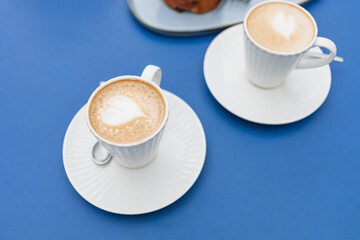 Close-up of latte coffee in beautiful, elegant white cups with pastries placed on a blue table.