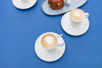 Close-up overhead view of cappuccino coffee in beautiful, elegant white cups with pastries placed on a blue table.