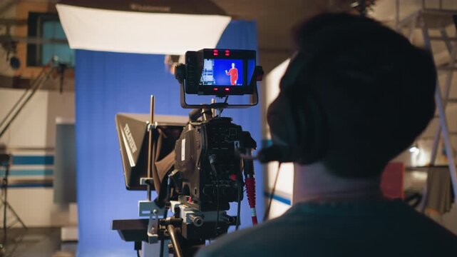 Cameraman wearing headset operating television camera with teleprompter during studio shoot while recording female presenter in red dress on blue screen background under professional lighting setup