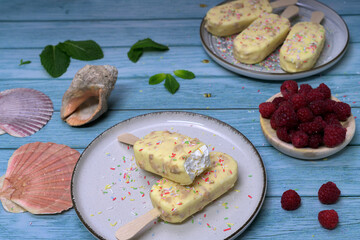 Homemade ice cream bars covered with white chocolate and red berries on a summery blue background, with raspberries and tropical decoration. Refreshing dessert concept