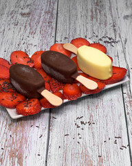 Top view of assorted chocolate ice cream bars on a bed of strawberries, surrounded by fruits and chocolate on rustic wooden background. Colorful and appetizing summer dessert