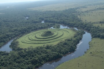 Aerial view of a circular, grassy mound in a rainforest, a river bend
