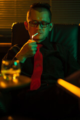Man sits on a sofa in the room, lighting a cigarette, holding a glass of whiskey, drinking with a frustrated expression on his face.