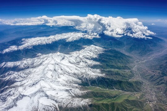 High-altitude view of snow-capped mountain ranges and valleys.  Vast landscape stretching across the horizon under a clear blue sky.  Large clouds over the peaks