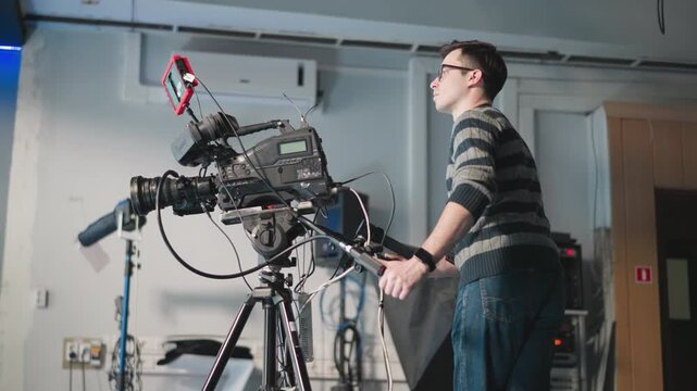 Man adjusting professional video camera on tripod in studio. Technician focused on camera setup with screen and equipment. Filming session ongoing in production environment
